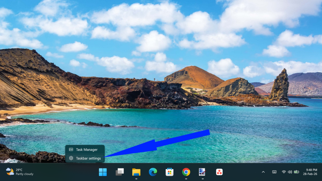 Windows desktop showing a blue arrow pointing to the Taskbar settings option in the taskbar right-click context menu.